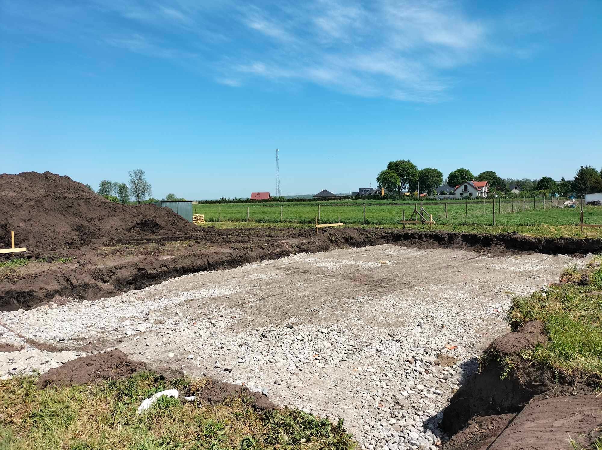 Przygotowany wykop pod fundamenty z wysypanym i wyrównanym kruszywem, obok hałda ziemi, w tle zielone pole i zabudowania pod błękitnym niebem.