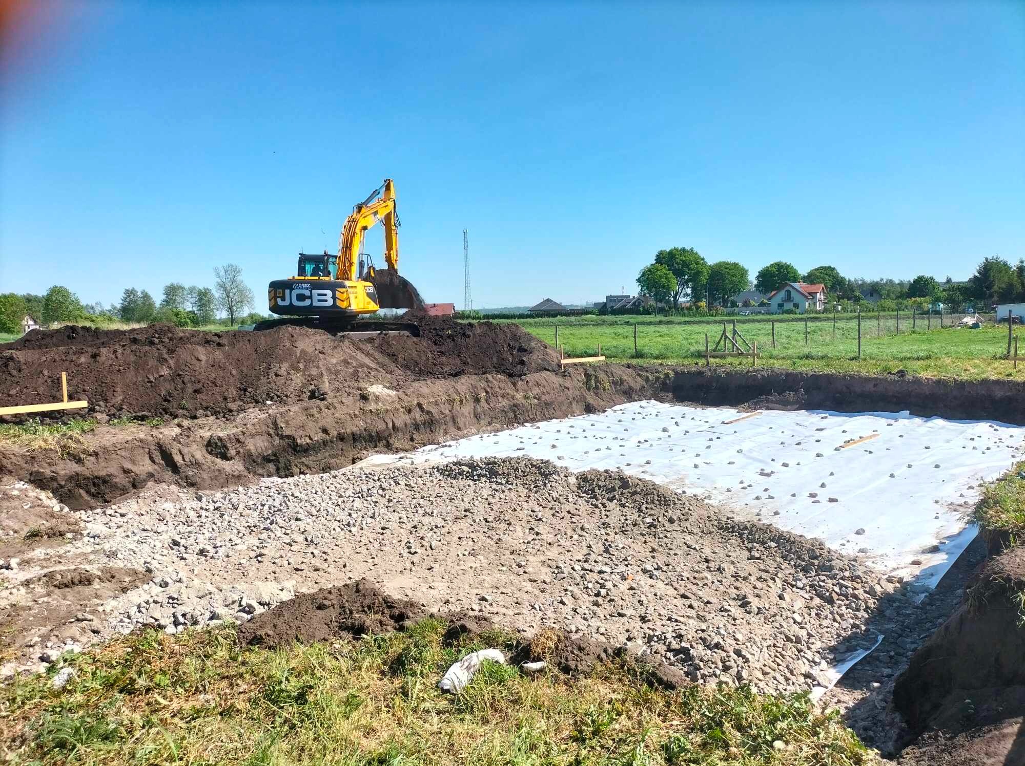 Żółta koparka JCB w trakcie prac ziemnych przy wykopie, widoczny żwir i biała geowłóknina, w tle zielone pola i zabudowania.