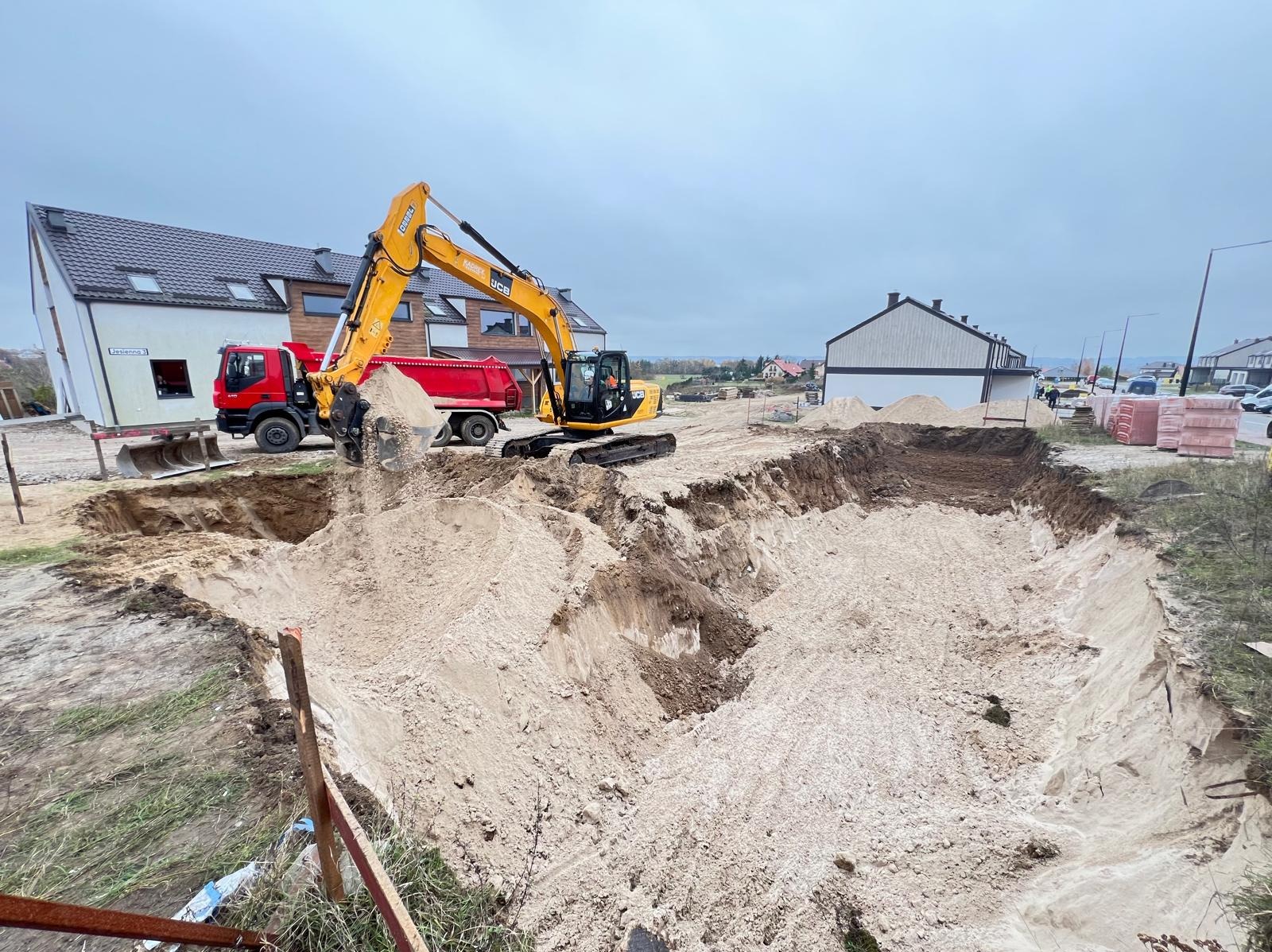 Żółta koparka JCB na placu budowy, wysypująca piasek do wykopu, obok stoi czerwona wywrotka, w tle zabudowa jednorodzinna, pochmurne niebo.