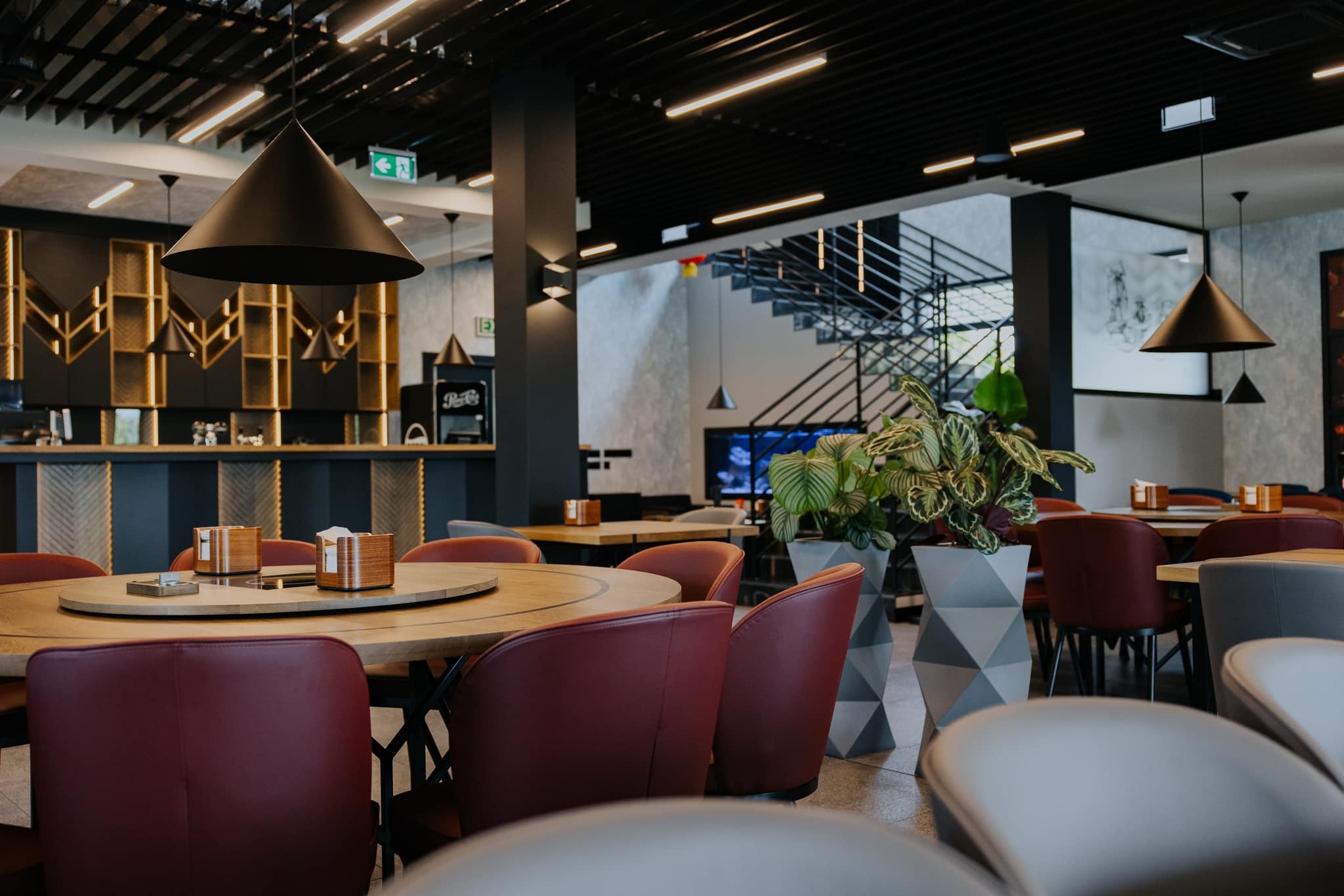 Wnętrze restauracji z okrągłymi stołami, bordowymi krzesłami, geometrycznymi donicami z roślinami, stołem barowym w tle i schodami na antresolę.