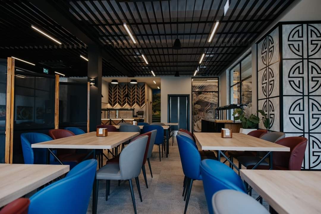 Nowoczesna aranżacja wnętrza restauracji z drewnianymi stołami, kolorowymi krzesłami i dekoracyjnym panelem ściennym z geometrycznym wzorem.