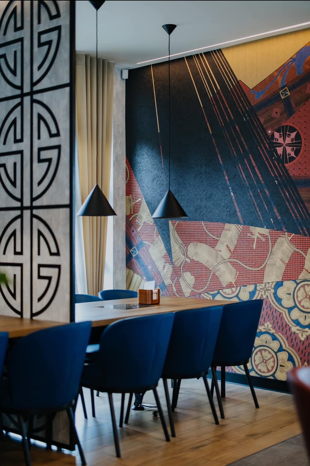 Stylowa aranżacja biura: Długi drewniany stół otoczony granatowymi krzesłami, nad nim wiszące czarne lampy. W tle wzorzysta tapeta i dekoracyjna ścianka działowa.