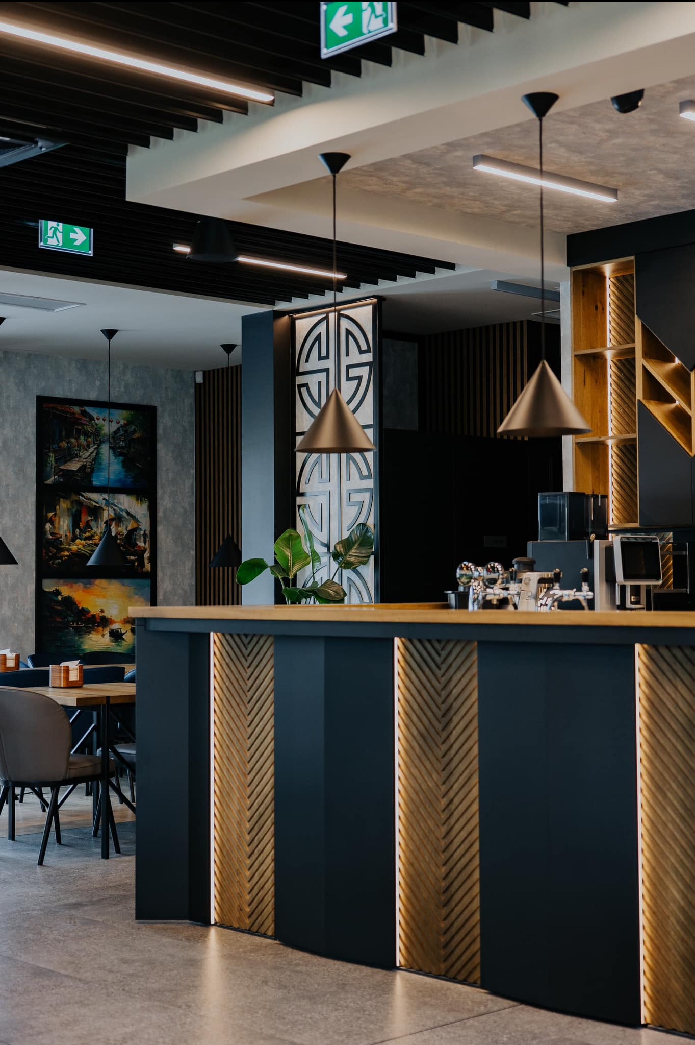 Nowoczesne wnętrze biurowe z ladą recepcyjną oświetloną ledami, stolikami i krzesłami. Na ścianie wisi obraz w trzech częściach. Widoczne lampy wiszące i dekoracyjne elementy.