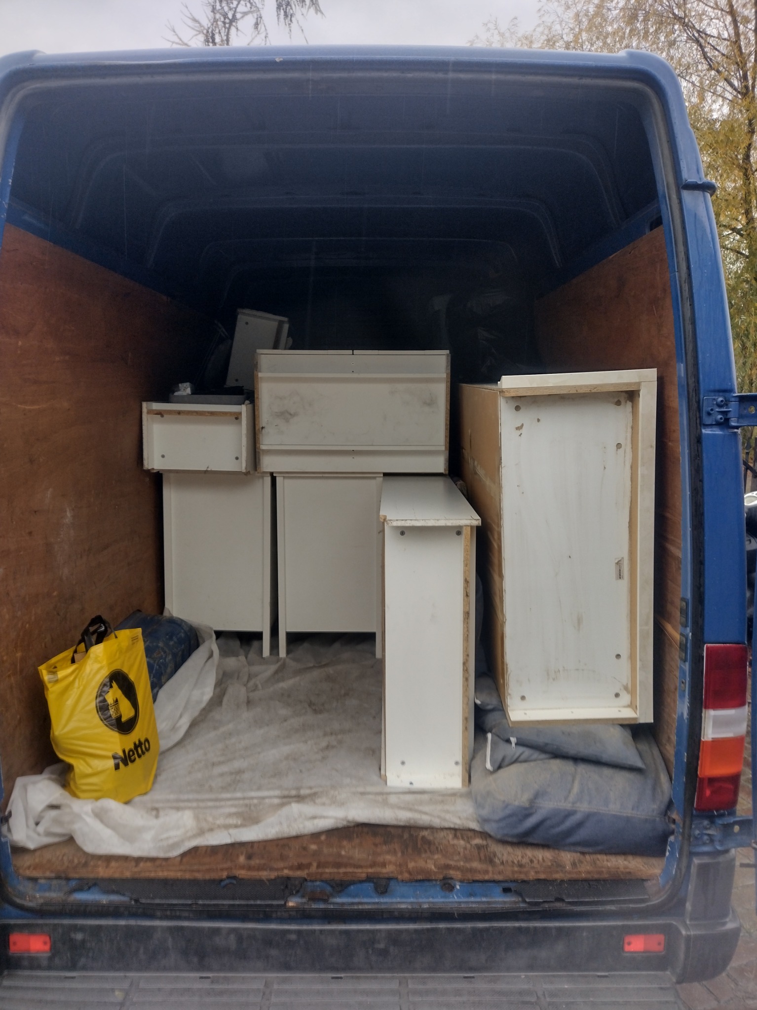Wnętrze niebieskiego busa transportowego wypełnione białymi meblami, zabezpieczonymi kocem i poduszkami. Żółta torba Netto w rogu. Widok z tyłu pojazdu.