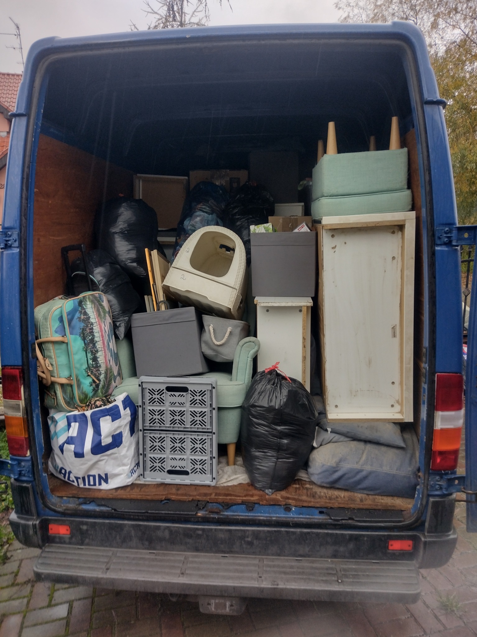 Załadunek mebli i rzeczy osobistych do niebieskiego busa. Widok z tyłu, otwarte drzwi. Wnętrze wypełnione po brzegi, widoczne torby, pudła, krzesła i inne przedmioty.