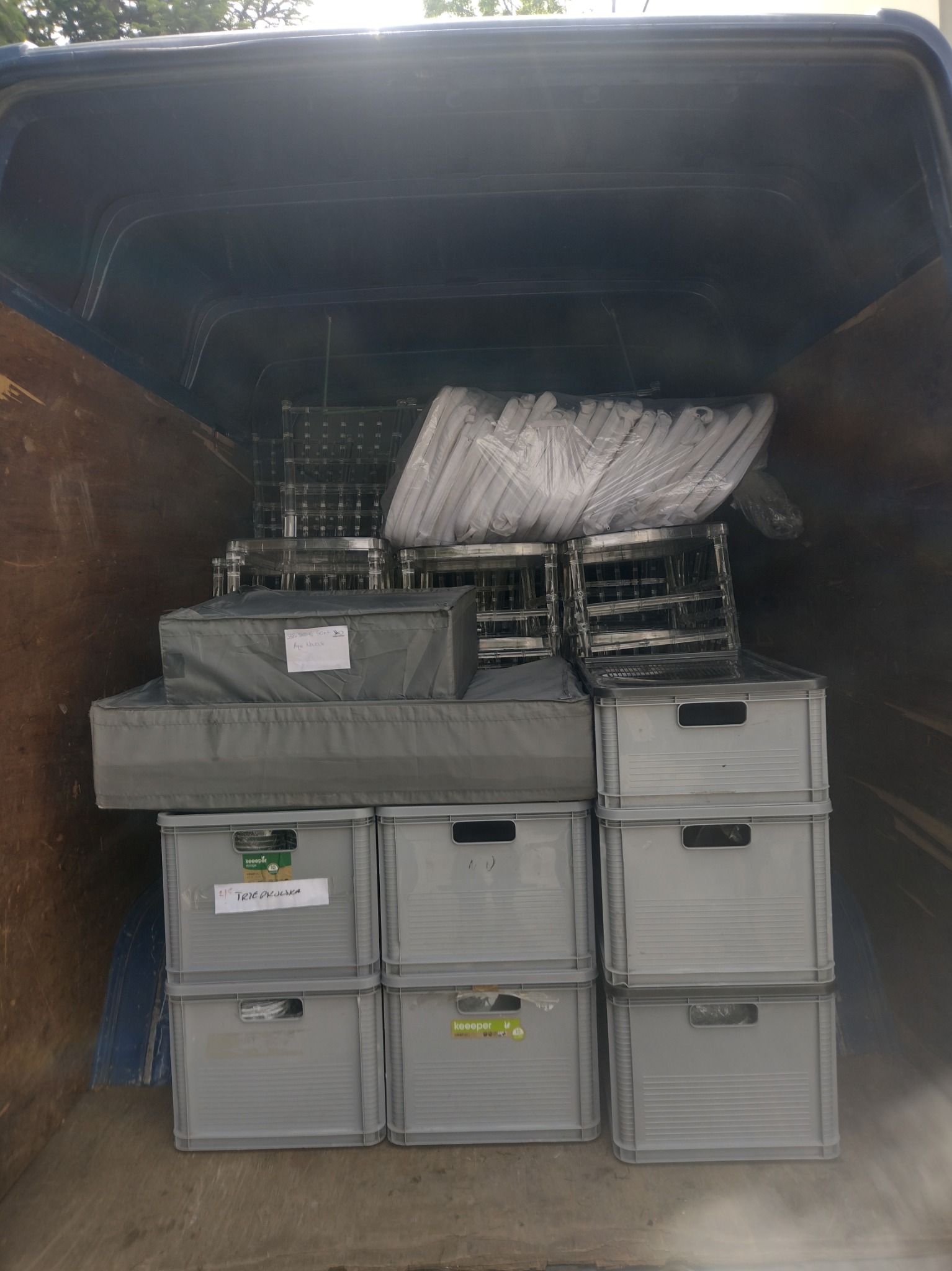 Wnętrze busa wypełnione szarymi skrzynkami, torbami i krzesłami w folii, gotowe do transportu. Widoczne etykiety adresowe na niektórych przedmiotach.