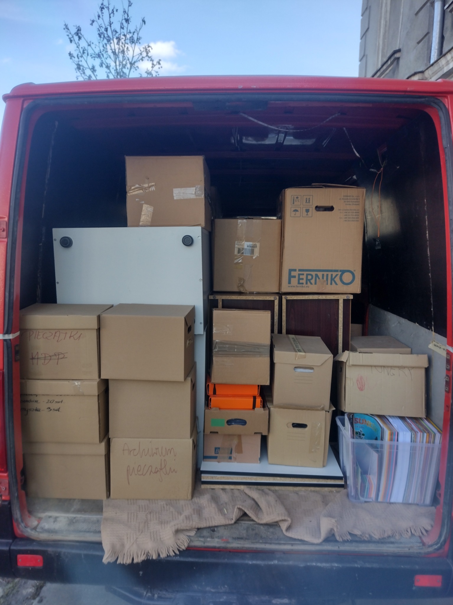 Czerwony bus załadowany kartonami z napisem 'Archiwum pieczątki', 'Ferniko' i białym regałem. Widok z tyłu pojazdu, przygotowanie do transportu.