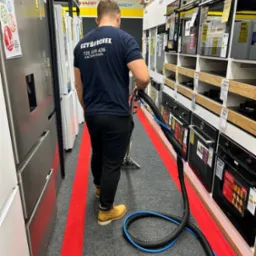 Mężczyzna w koszulce z napisem 'CZYŚCICIEL' czyści dywan odkurzaczem piorącym w sklepie z AGD, widoczne półki z piekarnikami i lodówkami.