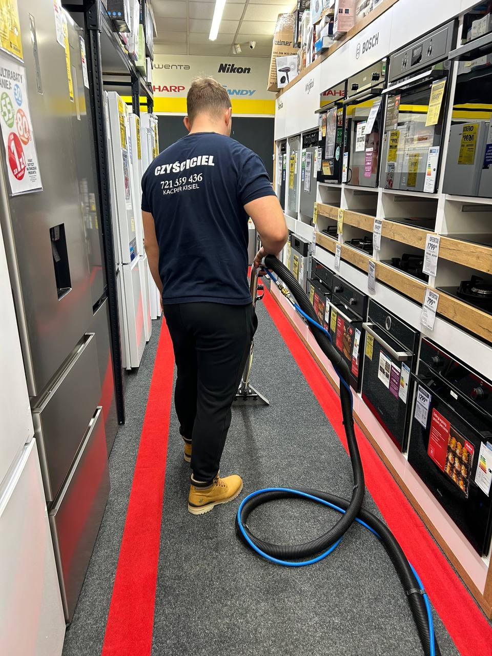 Mężczyzna w koszulce z napisem 'CZYŚCICIEL' czyści dywan odkurzaczem piorącym w sklepie z AGD, widoczne półki z piekarnikami i lodówkami.