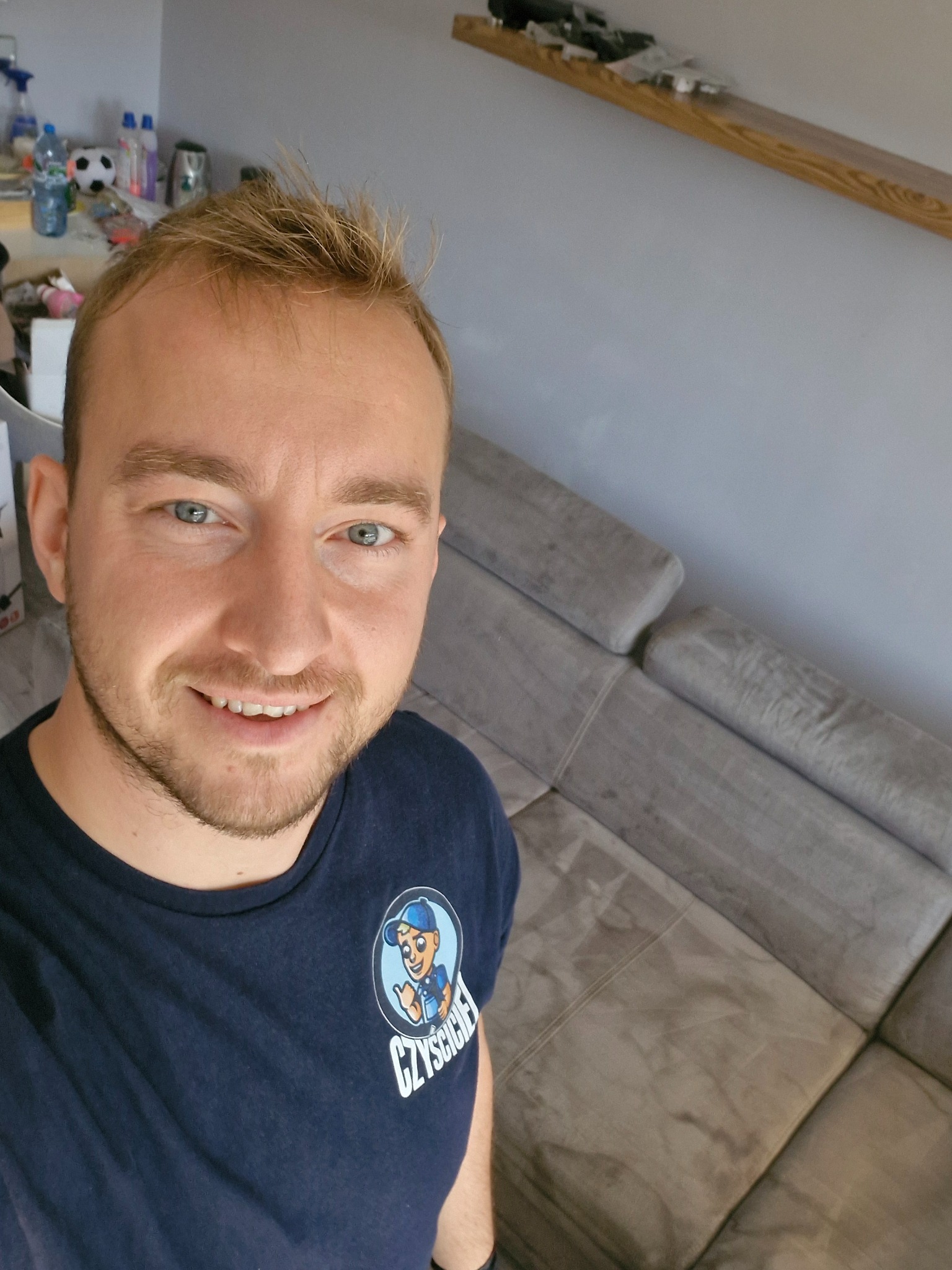 Uśmiechnięty mężczyzna w koszulce z logo firmy czyszczącej na tle szarej, modułowej kanapy w salonie.