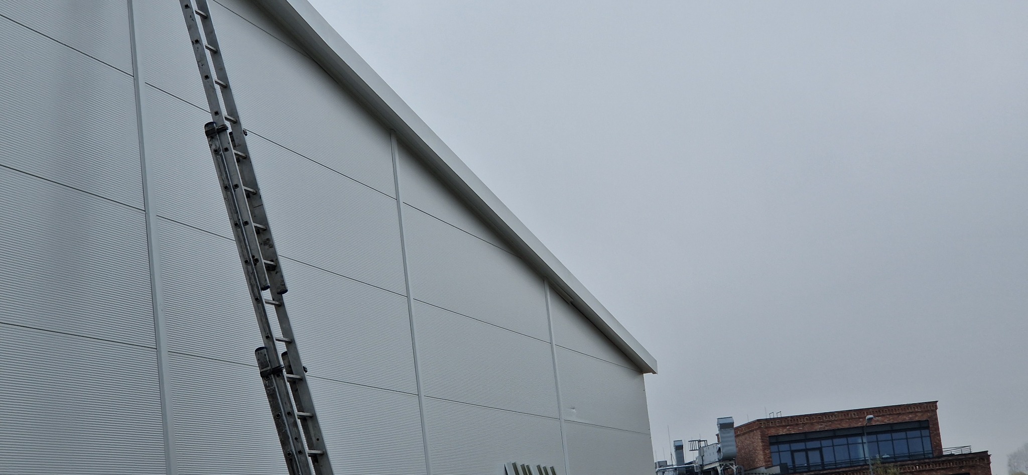Metalowa drabina oparta o ścianę hali przemysłowej z panelami, w tle widoczny budynek z czerwonej cegły i pochmurne niebo.