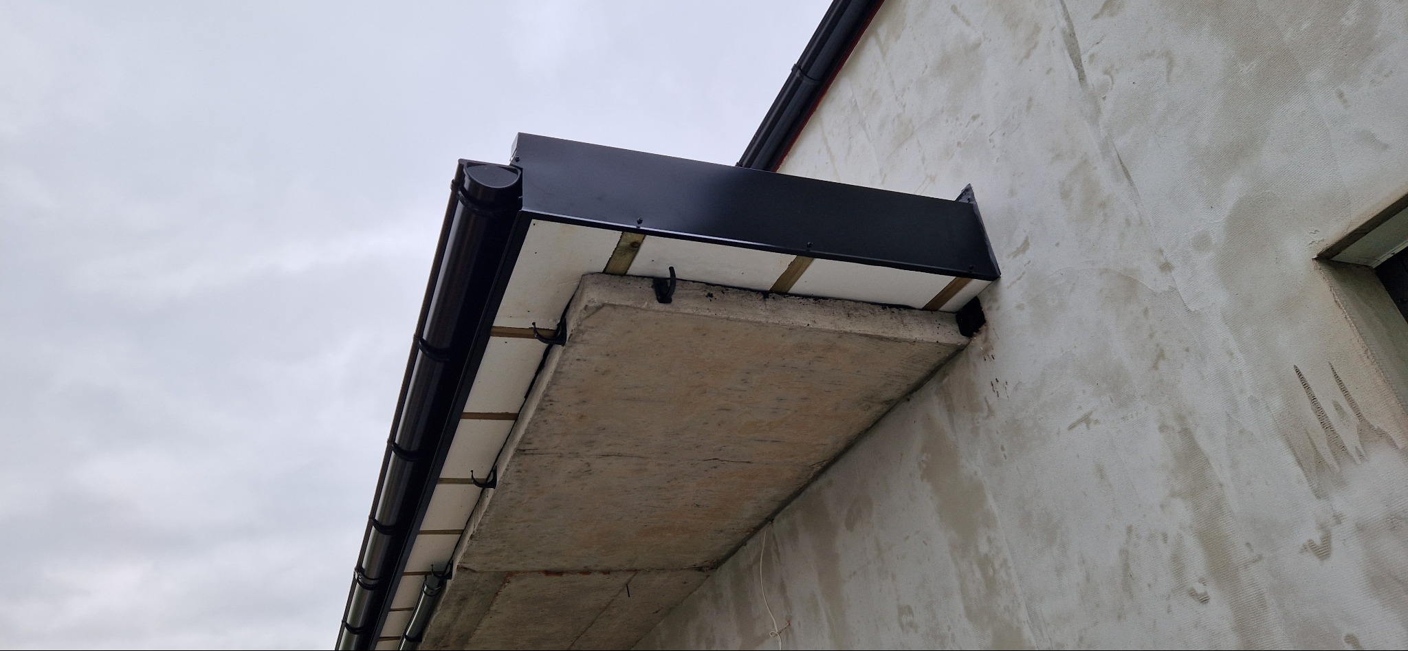 Zamontowana, ciemnobrązowa rynna dachowa z czarnym panelem maskującym, przymocowana do ściany budynku z widocznym betonowym elementem konstrukcyjnym.