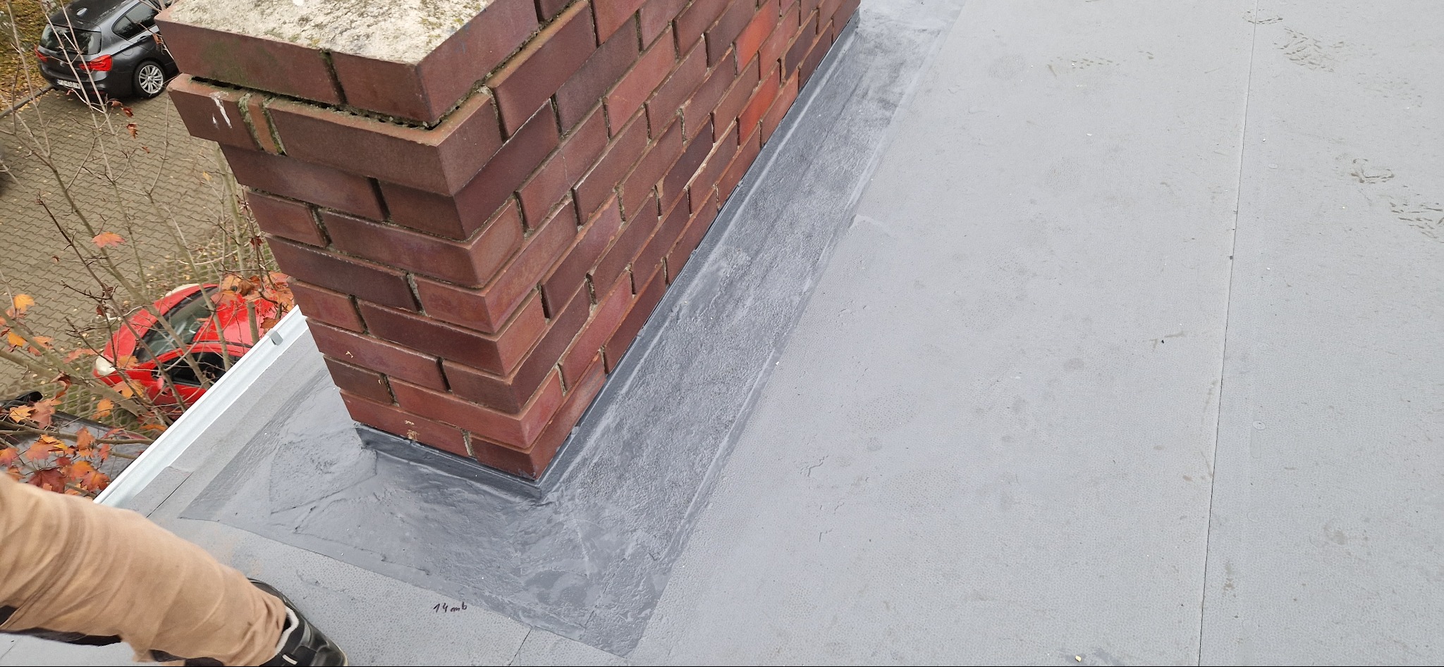 Szczegółowe uszczelnienie komina na płaskim dachu pokrytym szarą membraną, widoczne ślady butów dekarza i fragment czerwonego samochodu w tle.