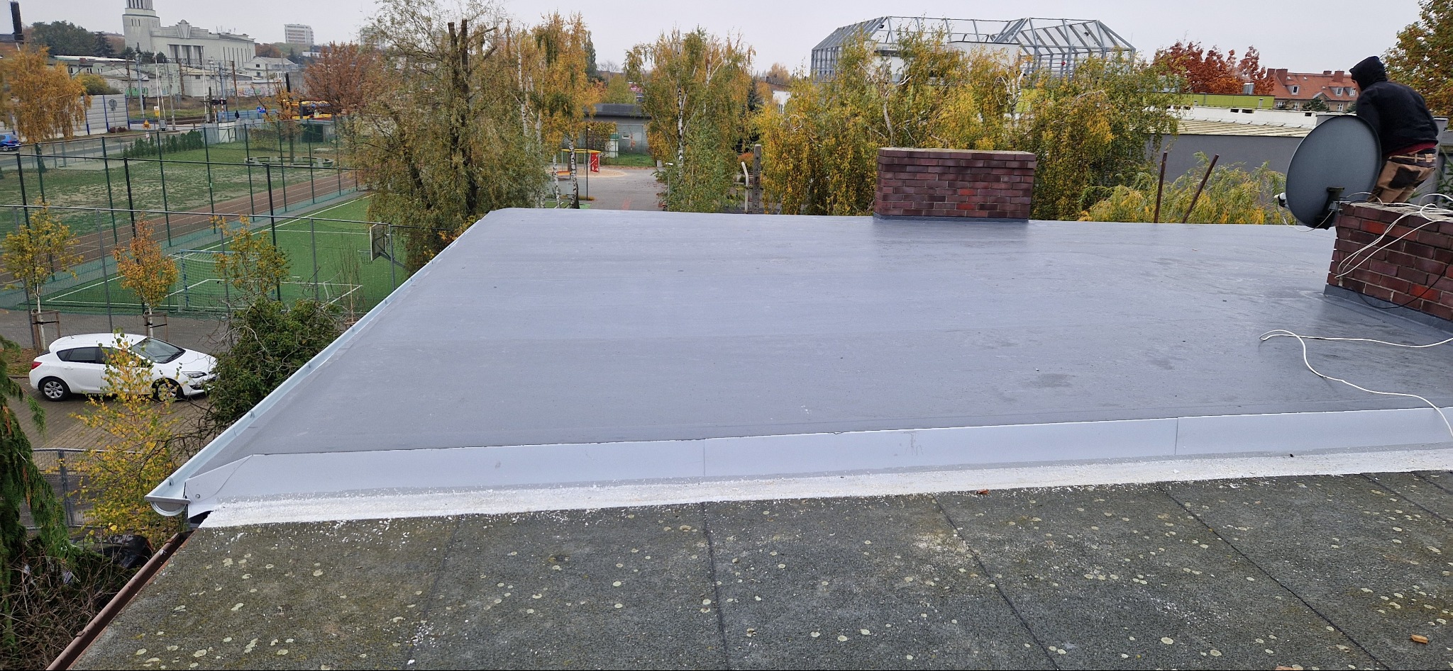 Szary, płaski dach pokryty papą termozgrzewalną z widoczną instalacją antenową na kominie z cegły i pracownikiem w czarnej bluzie. W tle drzewa i budynki.