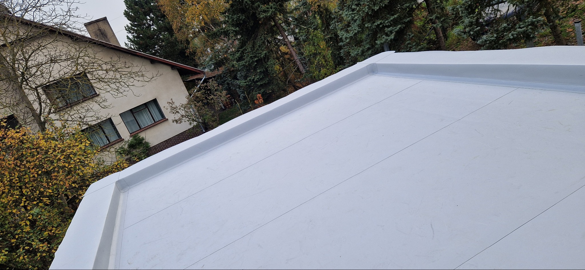 Biała, płaska powierzchnia dachu z widocznymi łączeniami i obróbkami blacharskimi, na tle drzew i budynku mieszkalnego