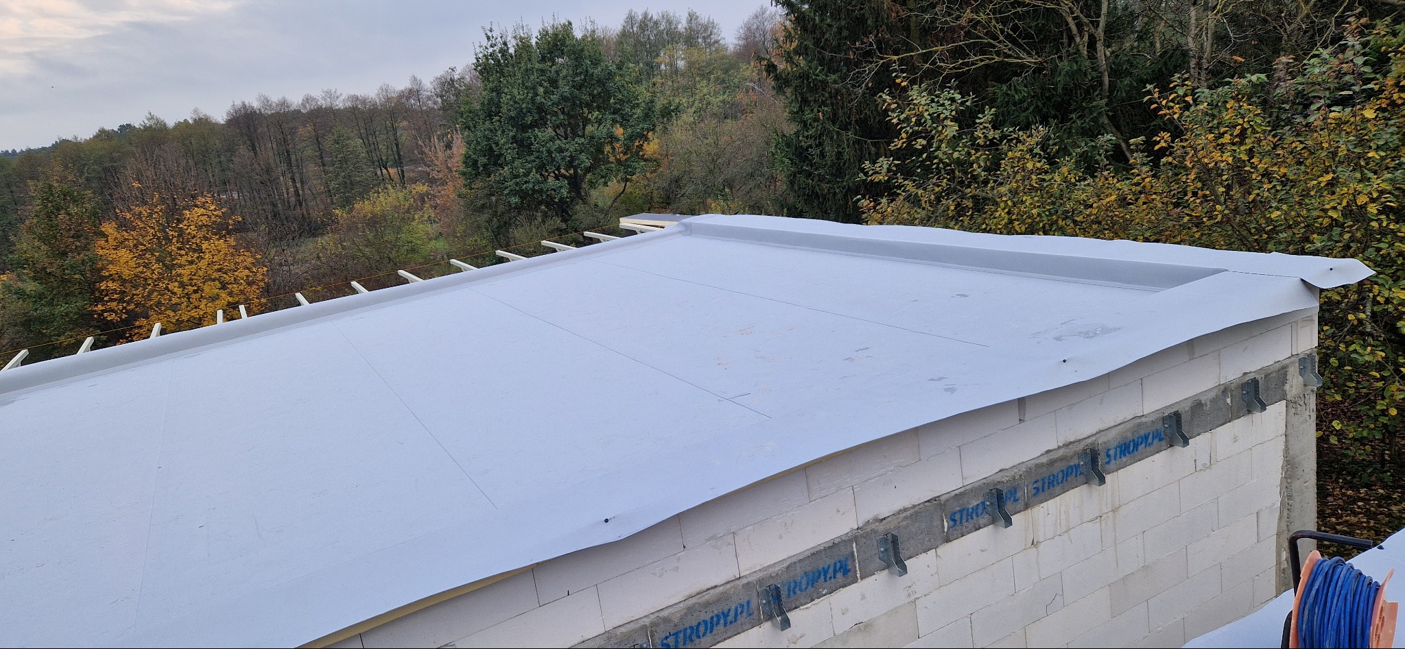 Płaski dach pokryty membraną PVC, widoczne mocowania na ścianach budynku z betonu komórkowego, las w tle.