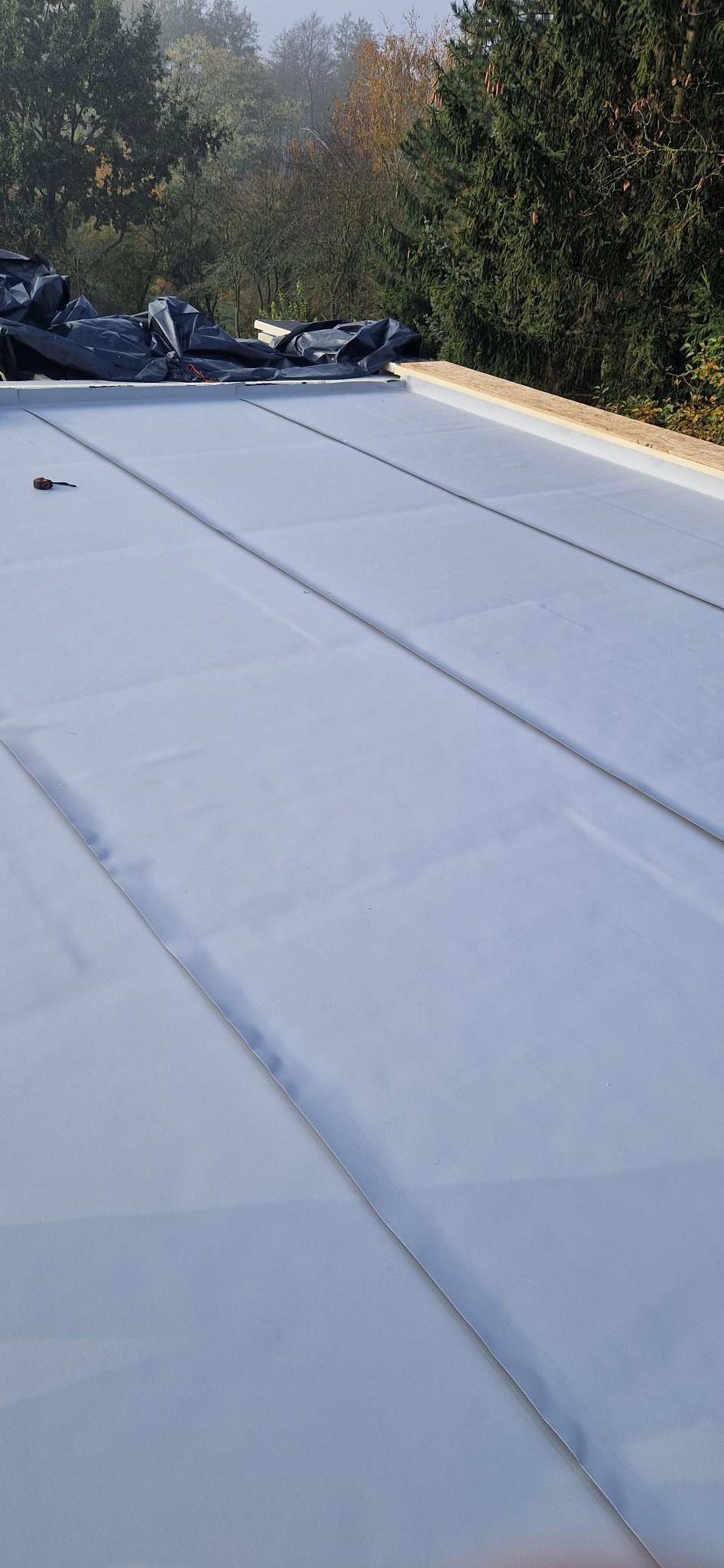 Szary dach z blachy na drewnianej konstrukcji, w tle drzewa i czarna folia ochronna.
