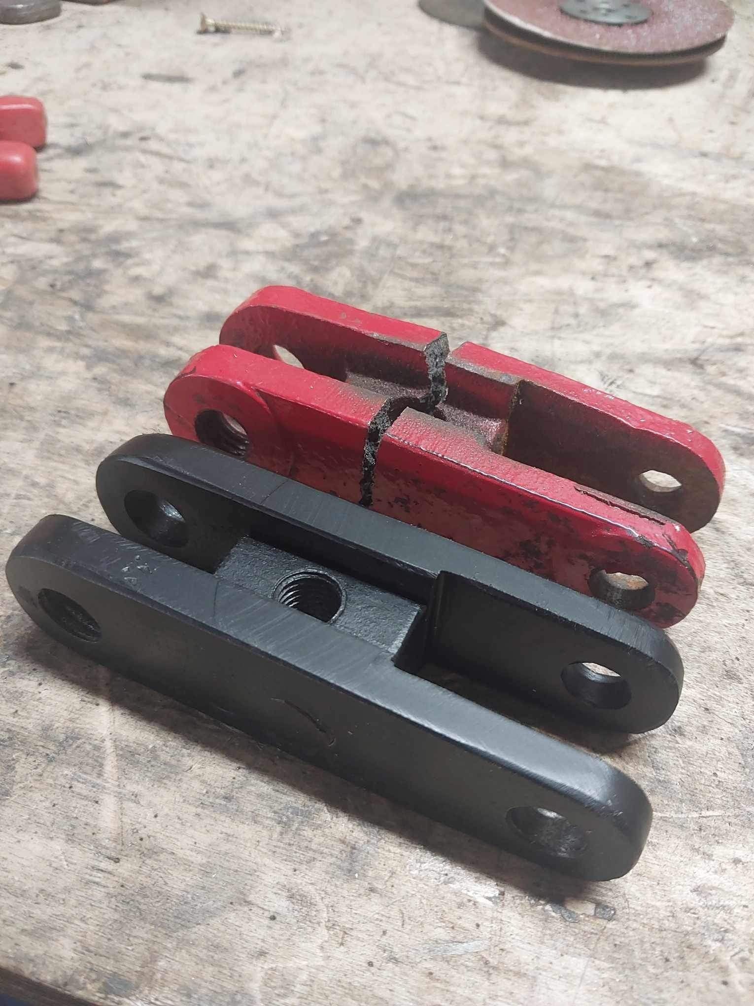 Dwa czerwone i jeden czarny, metalowe łączniki z otworami i gwintem, leżące na brudnym, drewnianym stole warsztatowym. Jeden z czerwonych łączników jest pęknięty.