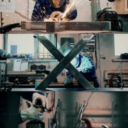 Mira Media - Spawanie metalowej konstrukcji w kształcie litery X w warsztacie, widoczne iskry, spawacz w ochronnej masce i kombinezonie, w tle narzędzia i wyposażenie warsztatu.