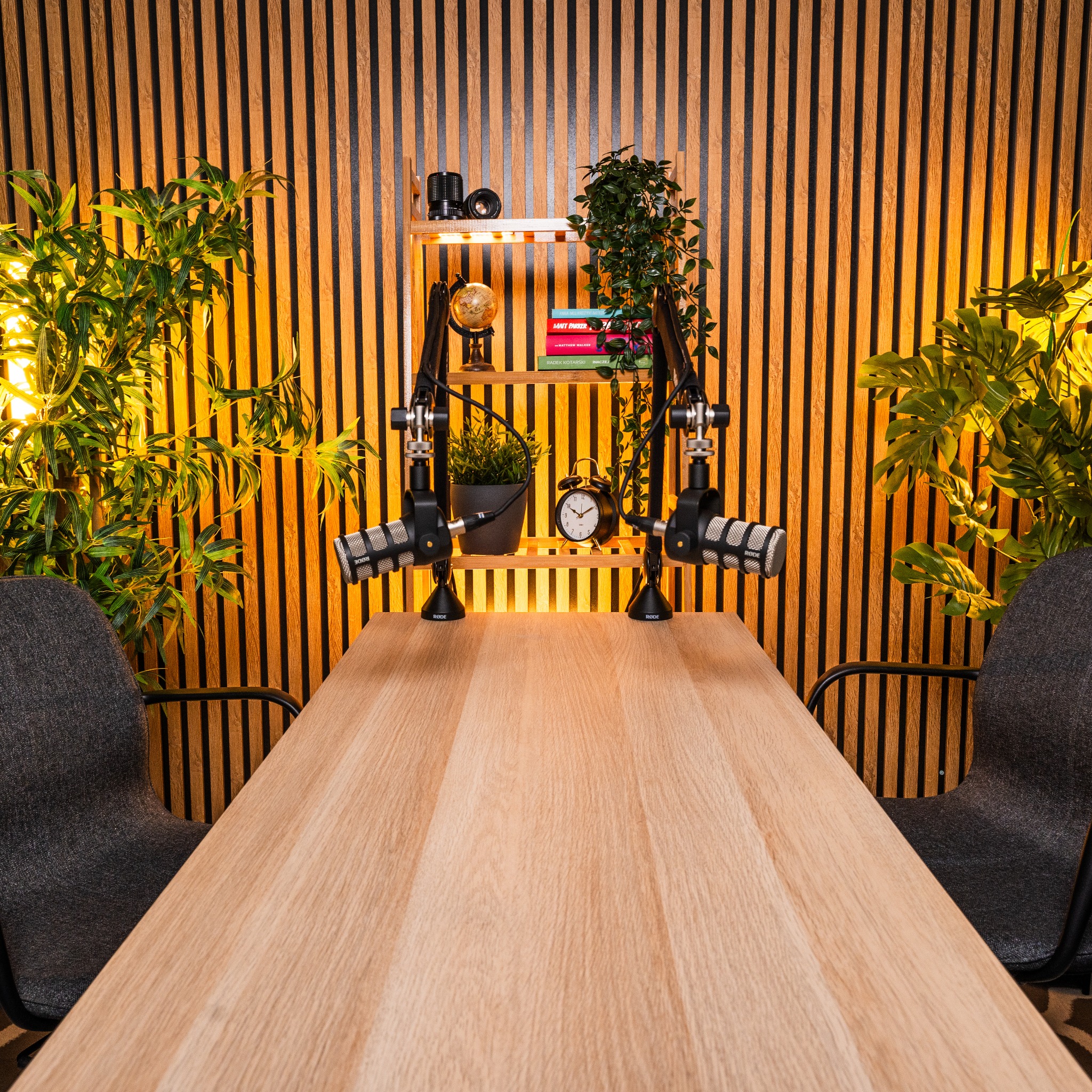 Profesjonalne studio nagraniowe z dwoma mikrofonami Rode na statywach, drewnianym biurkiem, krzesłami, półką z książkami, globusem i roślinami na tle drewnianej ściany z lameli.