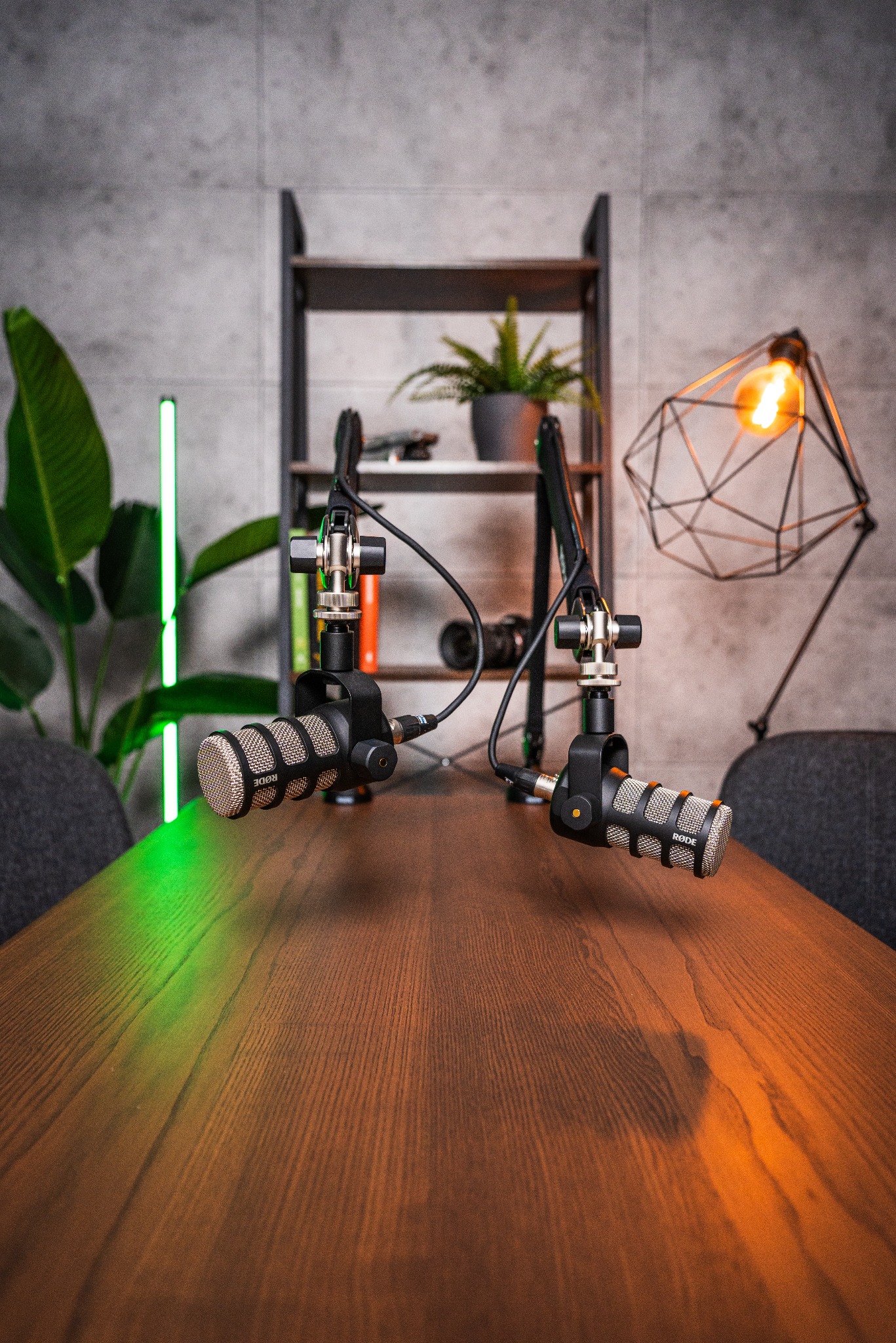 Dwa mikrofony Rode na wysięgnikach stoją na drewnianym stole, oświetlone zielonym i pomarańczowym światłem, w tle regał z roślinami i lampa w kształcie diamentu.