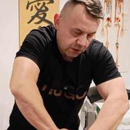 BOGDAN POCHCIOŁ NATURMED - Mężczyzna w czarnej koszulce wykonuje masaż pleców pacjentki; w tle plakat z układem kostnym i dekoracja ścienna z chińskim znakiem.