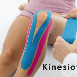 BOGDAN POCHCIOŁ NATURMED - Nakładanie elastycznych taśm kinezjologicznych w kolorach niebieskim i różowym na kolano pacjenta przez osobę w białym fartuchu medycznym.