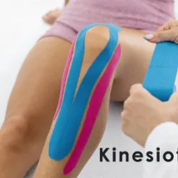 Nakładanie elastycznych taśm kinezjologicznych w kolorach niebieskim i różowym na kolano pacjenta przez osobę w białym fartuchu medycznym.