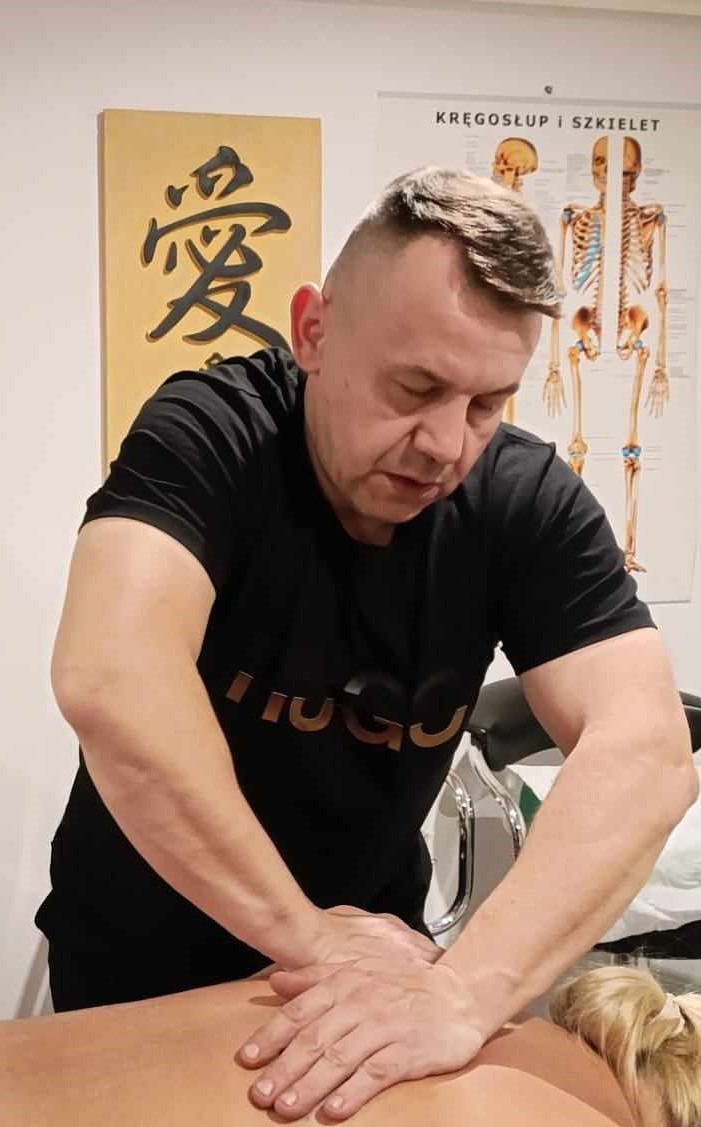 Mężczyzna w czarnej koszulce wykonuje masaż pleców pacjentki; w tle plakat z układem kostnym i dekoracja ścienna z chińskim znakiem.