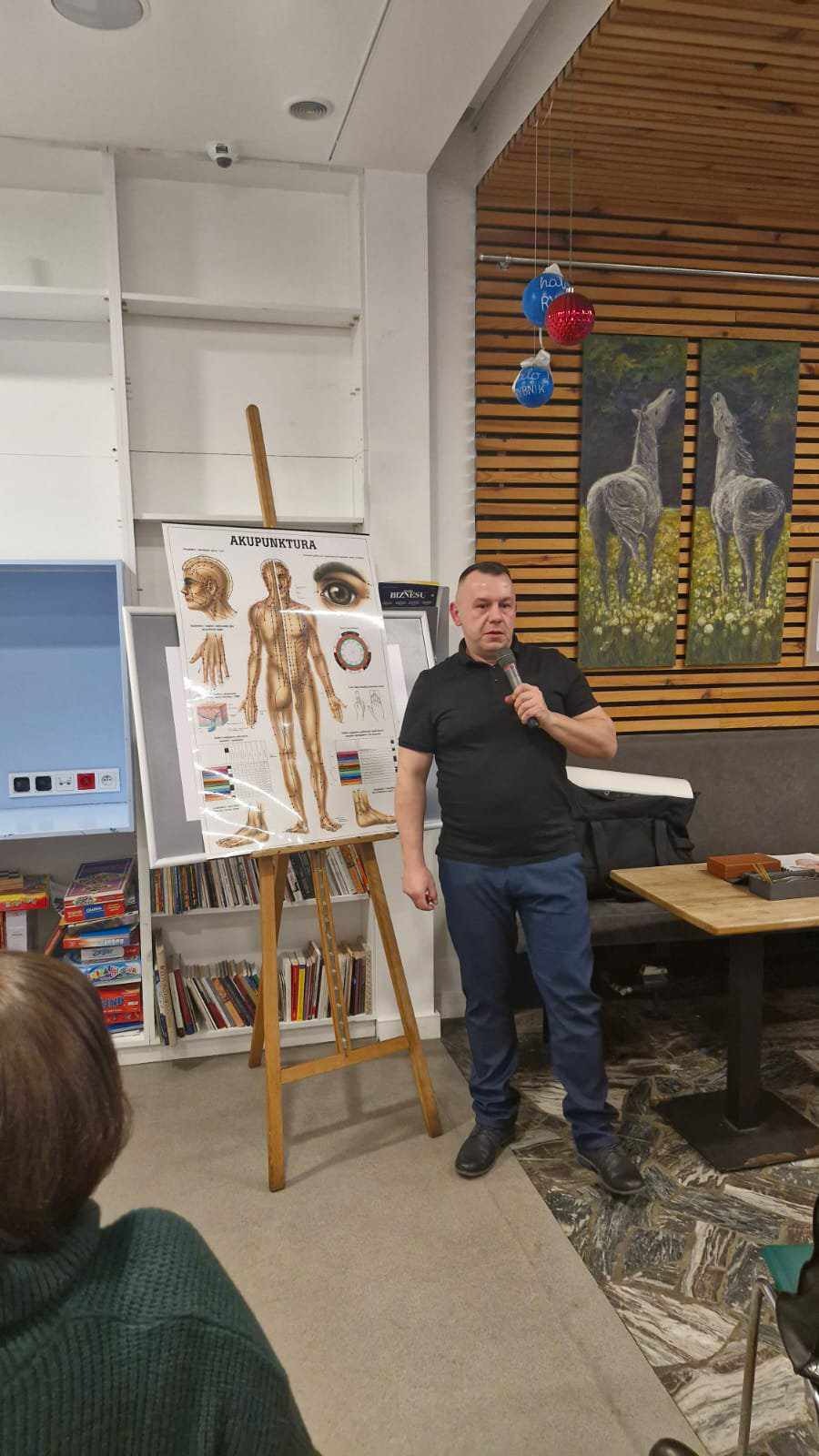 Mężczyzna w czarnej koszulce prezentuje plakat z mapą punktów akupunktury na sztaludze, trzymając mikrofon, w tle regał z książkami i obraz z końmi.