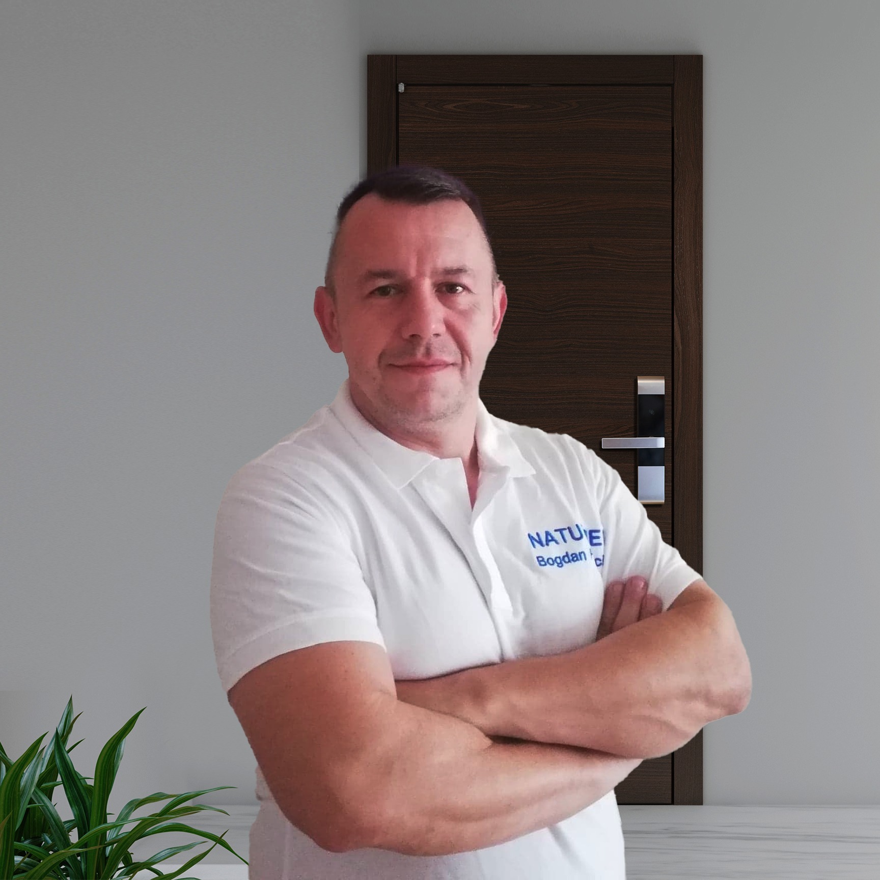 Portret mężczyzny w białej koszulce polo z logo firmy 'Nature Bogdan', stojącego na tle szarej ściany i brązowych drzwi z elektronicznym zamkiem.