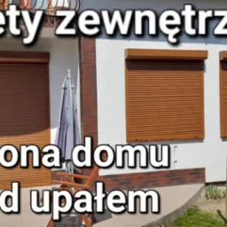 Brązowe rolety zewnętrzne zamontowane na fasadzie domu z białym tynkiem i ceglanym cokołem, widoczne ogrodzenie i parasol ogrodowy.