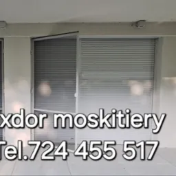 Zamontowane moskitiery ramkowe w kolorze białym na tarasie, częściowo zasłonięte opuszczonymi roletami zewnętrznymi, widoczny numer telefonu firmy Exdor.