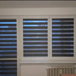 Trzy okna z szarymi roletami dzień noc, częściowo zasłonięte. Widoczne budynki za oknem. Na dole grzejnik.