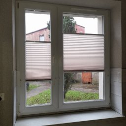 Exdor rolety żaluzje plisy moskitiery - Okno z zamontowanymi jasnymi plisami, widok na zewnątrz z zielenią i budynkiem w tle. Plisy częściowo zasłonięte, na parapecie grzejnik.