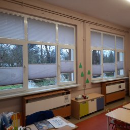 Exdor rolety żaluzje plisy moskitiery - Dwa okna z zamontowanymi plisami w jasnym pomieszczeniu. Widok z okna na drzewa. Plisy częściowo zasłonięte, z widoczną strukturą materiału.