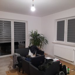 Exdor rolety żaluzje plisy moskitiery - Wnętrze pokoju z szarymi roletami dzień noc na oknach i drzwiach balkonowych, widoczne meble ogrodowe i roślina doniczkowa. Oświetlenie sufitowe.