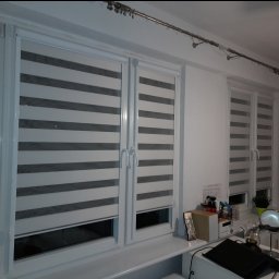 Exdor rolety żaluzje plisy moskitiery - Dwa okna z roletami dzień-noc w szaro-białe pasy, zamontowane w białych ramach okiennych. Na parapecie różne przedmioty, w tle trzecie okno z podobną roletą.