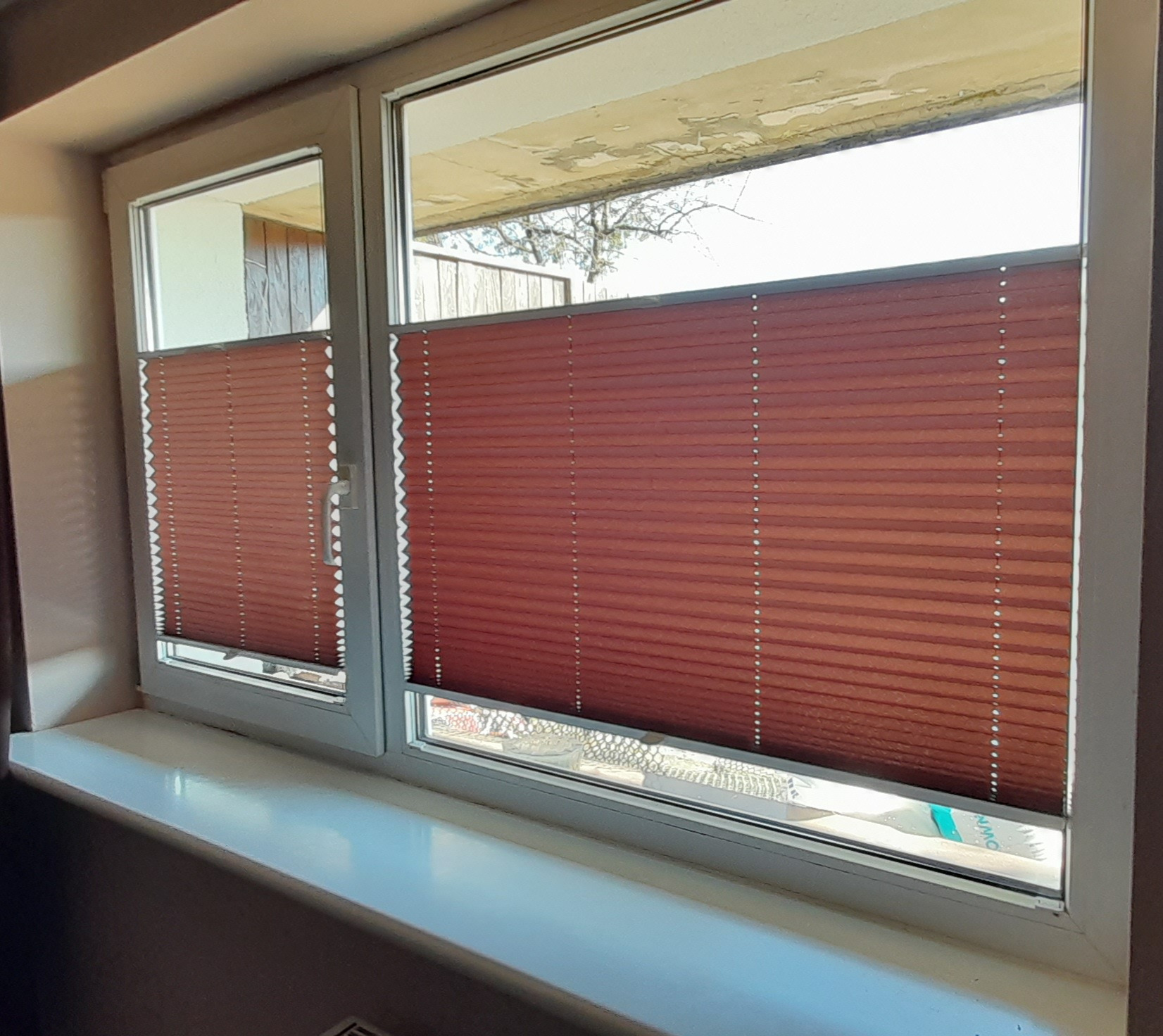 Zamknięte, brązowe plisy okienne w białej ramie okiennej, widok z wnętrza, na parapecie leży biało-turkusowy przedmiot.