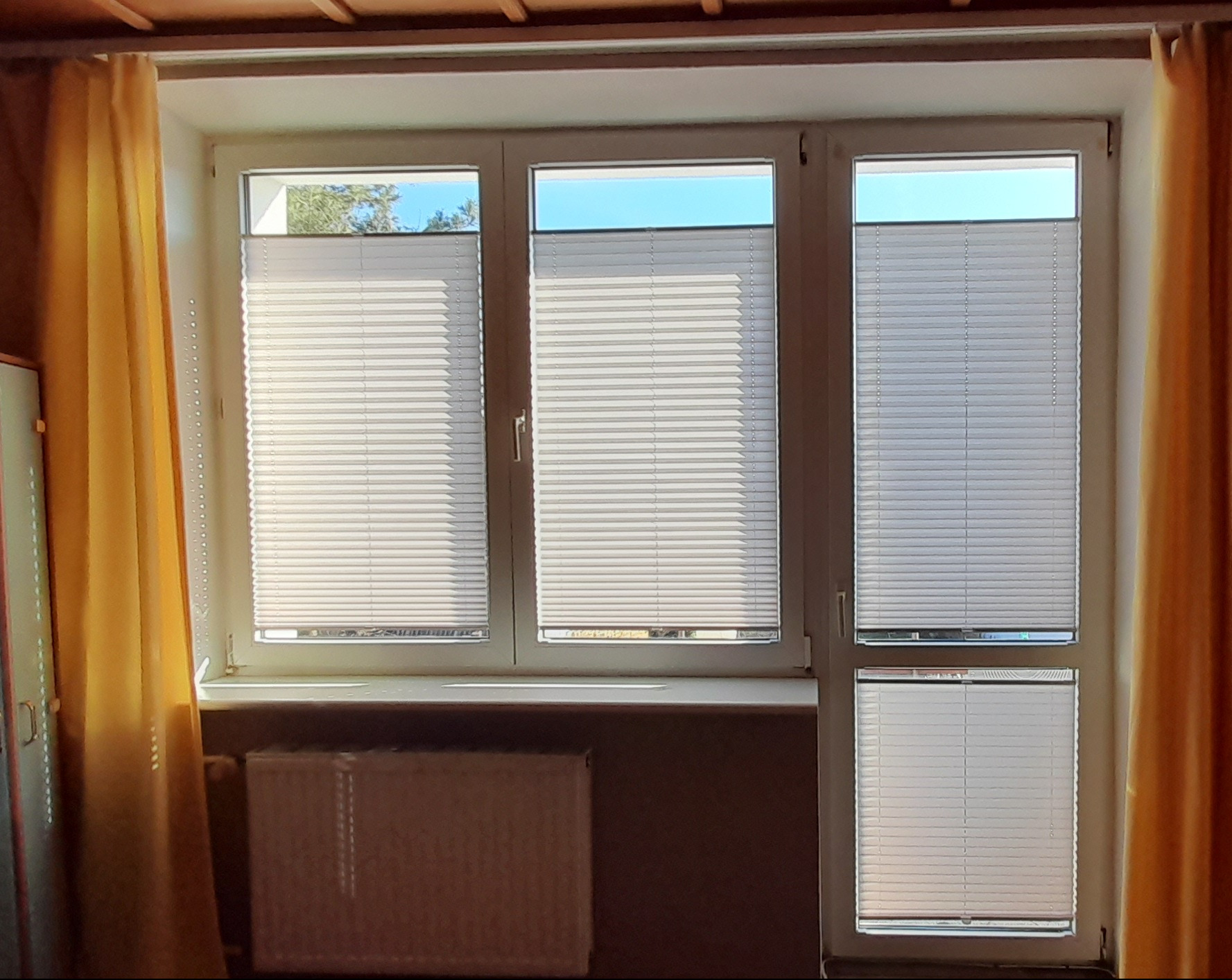 Trzy okna z białymi plisami, częściowo zasłonięte żółtymi zasłonami, z widocznym grzejnikiem pod parapetem.