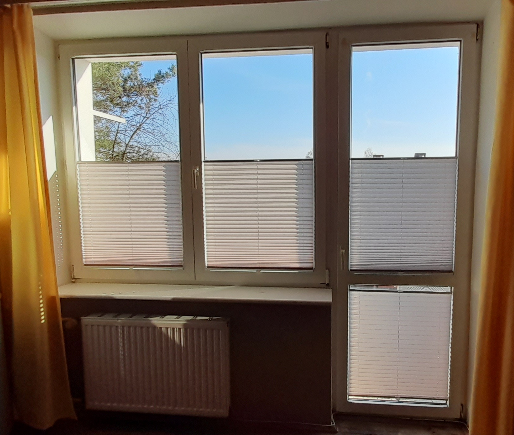 Trzyczęściowe okno z białymi ramami, wyposażone w jasne, poziome przesłony okienne, częściowo zasłonięte, widok na błękitne niebo i drzewa, żółte zasłony po bokach okna, grzejnik pod oknem.