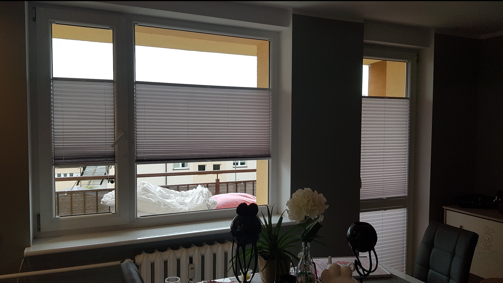 Szare plisy okienne zamontowane w oknach mieszkania z widokiem na balkon. Częściowo zasłonięte, z widocznymi elementami dekoracyjnymi na parapecie.