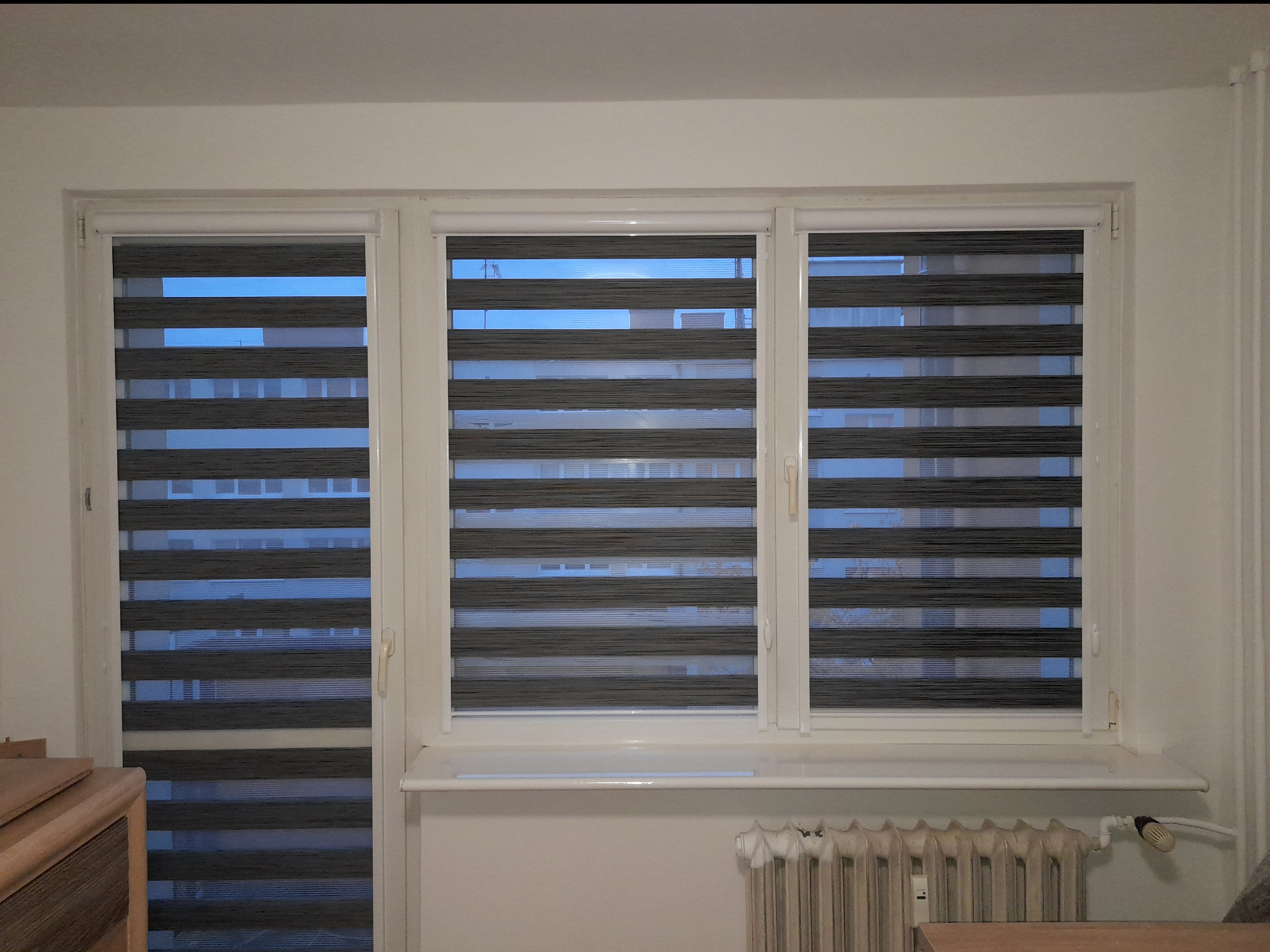 Trzy okna z szarymi roletami dzień noc, częściowo zasłonięte. Widoczne budynki za oknem. Na dole grzejnik.