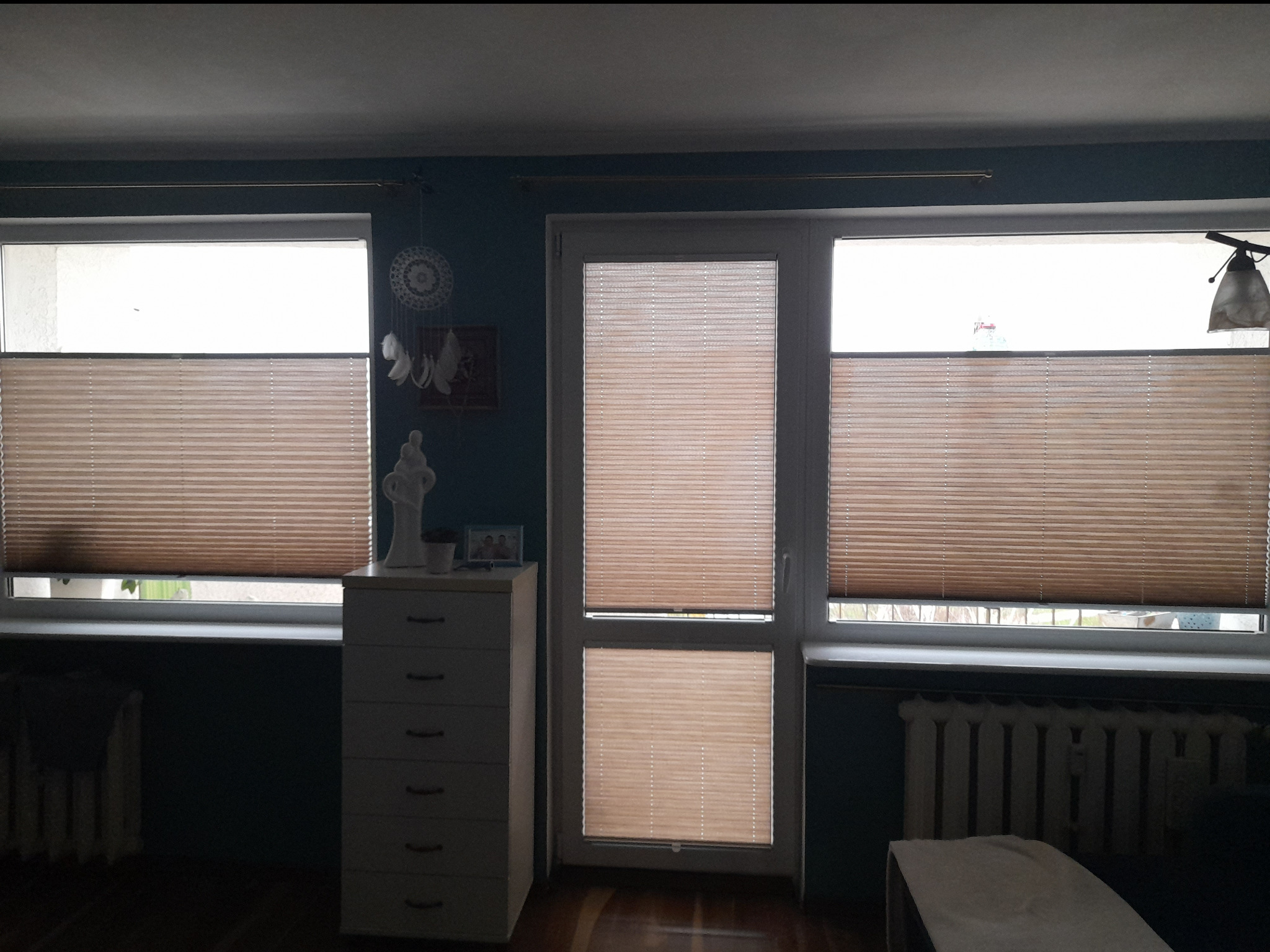 Beżowe plisy zamontowane w oknach i drzwiach balkonowych, widoczne z wnętrza pokoju o ciemnych ścianach. Na parapecie dekoracje, obok komoda.