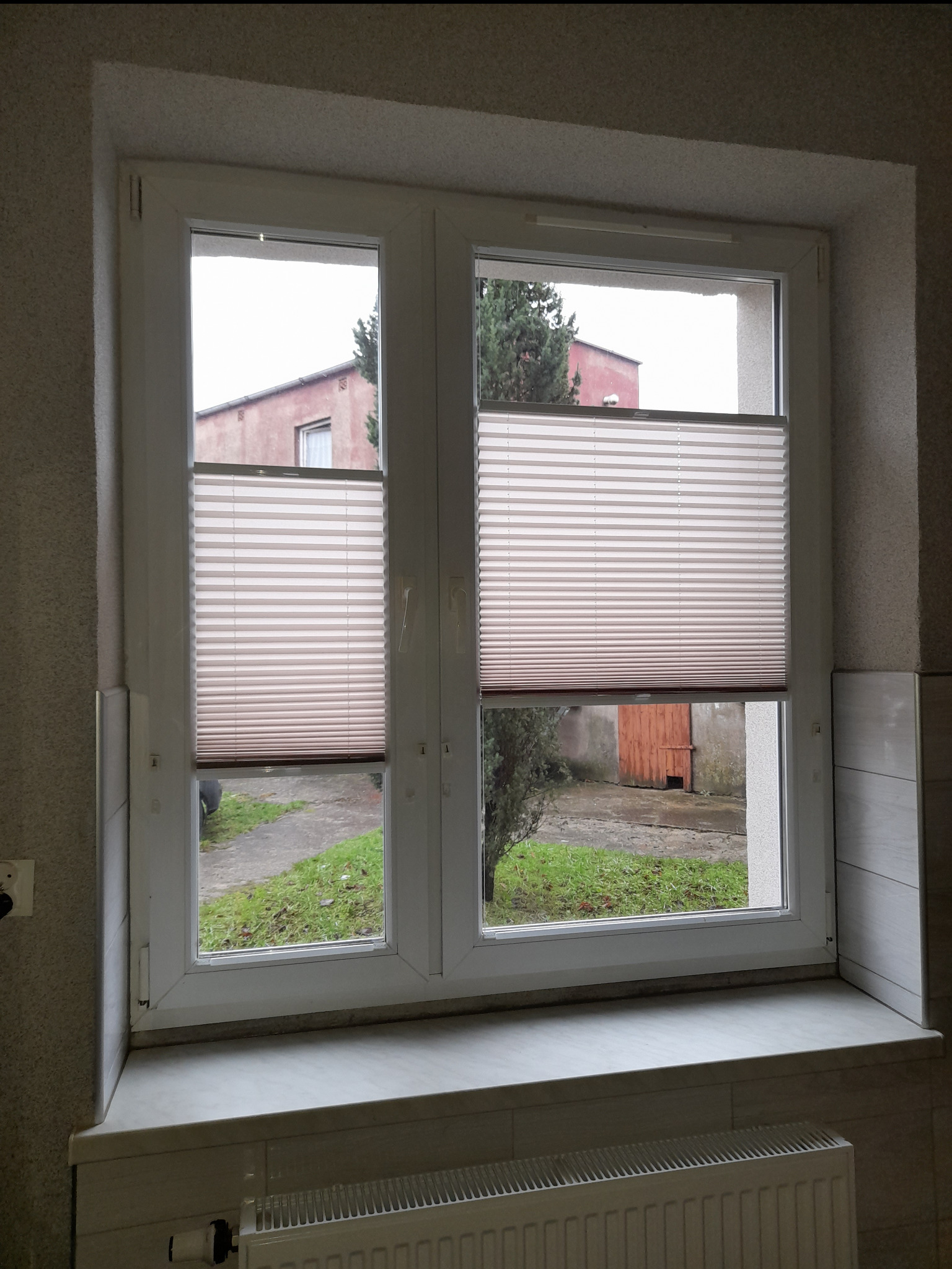 Okno z zamontowanymi jasnymi plisami, widok na zewnątrz z zielenią i budynkiem w tle. Plisy częściowo zasłonięte, na parapecie grzejnik.