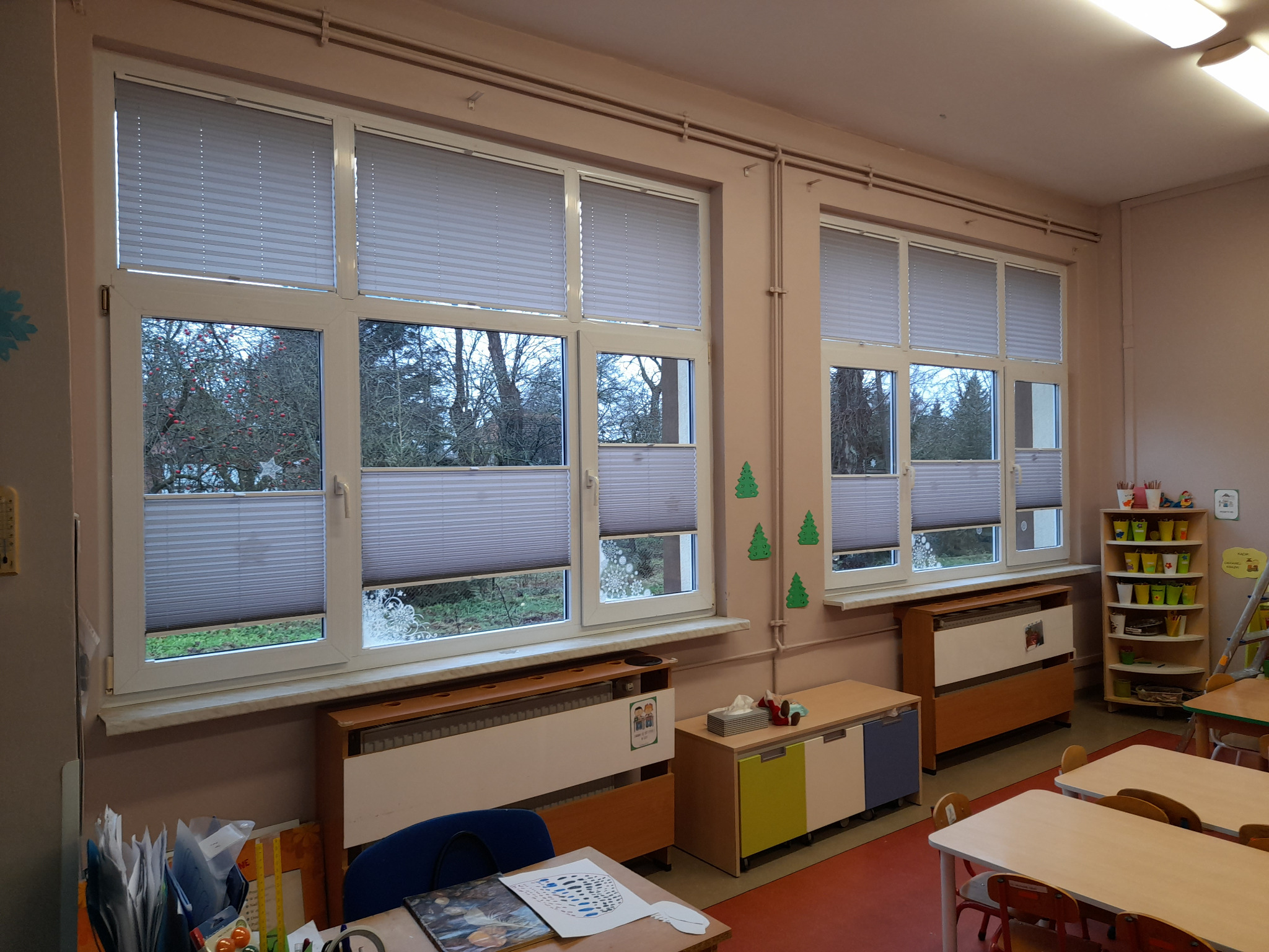 Dwa okna z zamontowanymi plisami w jasnym pomieszczeniu. Widok z okna na drzewa. Plisy częściowo zasłonięte, z widoczną strukturą materiału.