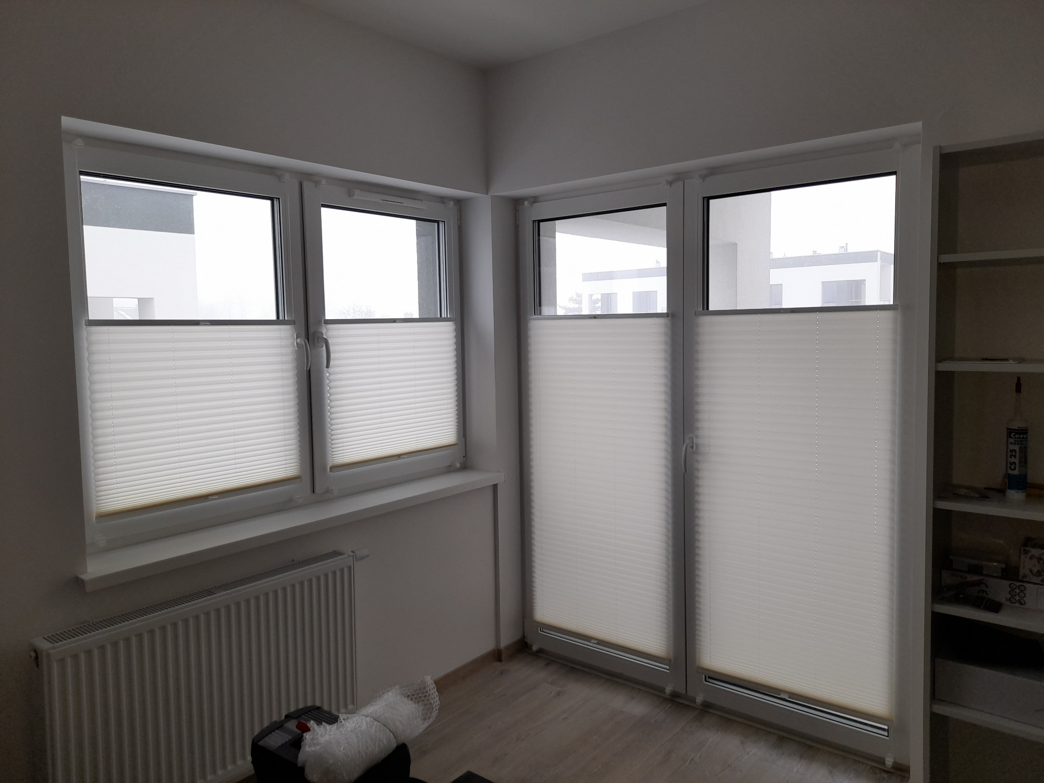 Jasne plisy okienne zamontowane w oknach w białych ramach, widok na budynki z zewnątrz. Grzejnik i regał w tle.