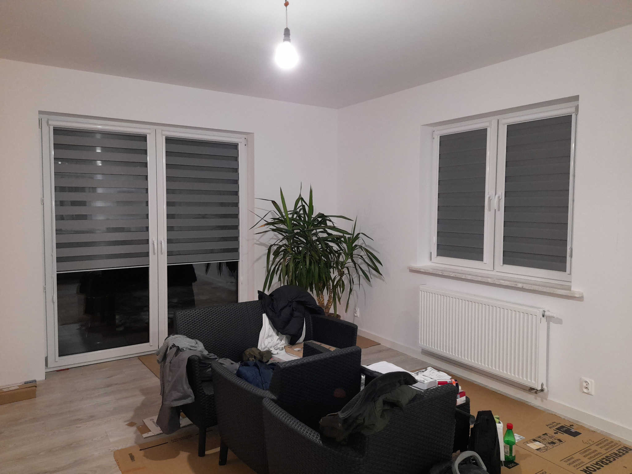 Wnętrze pokoju z szarymi roletami dzień noc na oknach i drzwiach balkonowych, widoczne meble ogrodowe i roślina doniczkowa. Oświetlenie sufitowe.