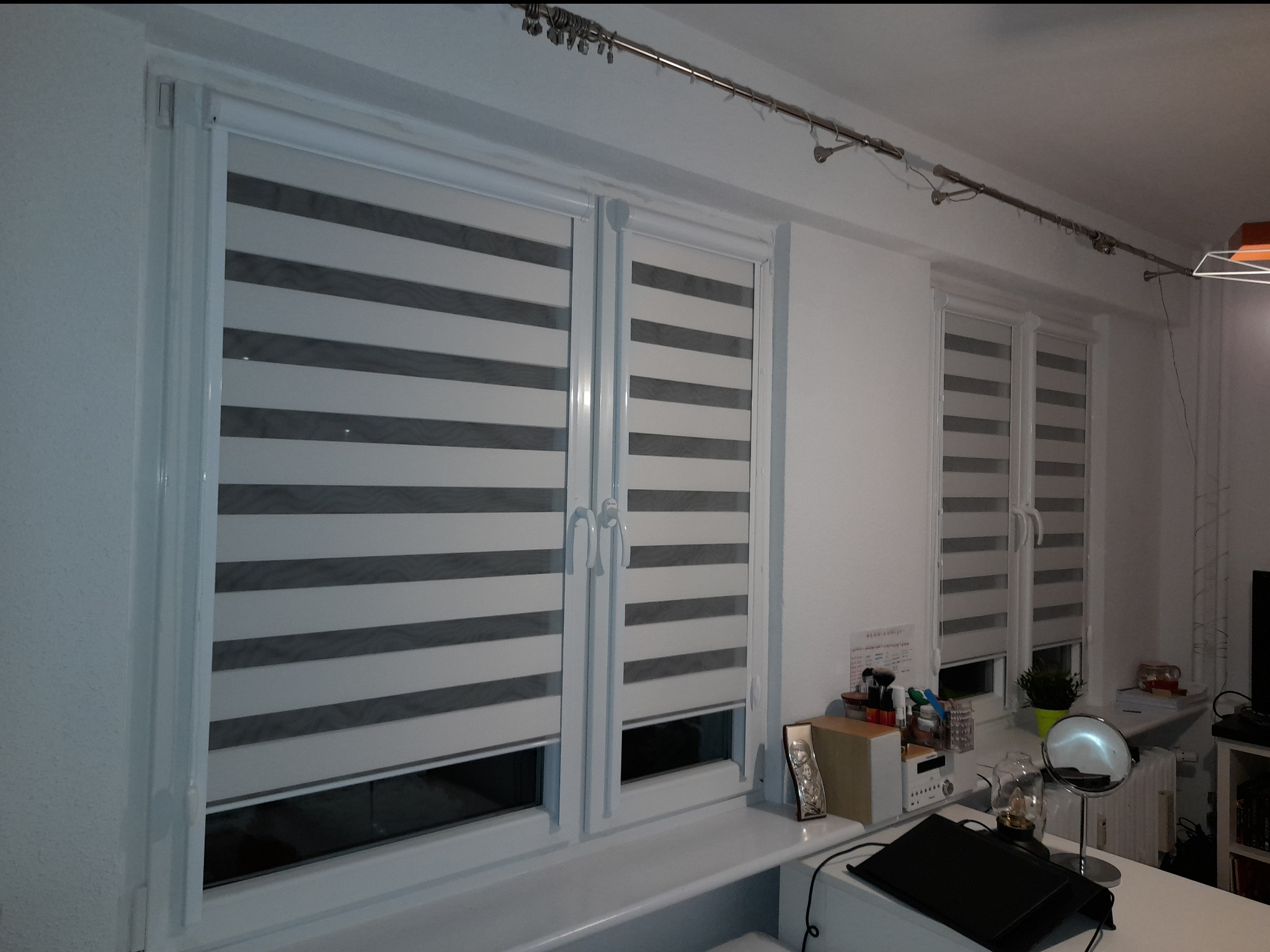 Dwa okna z roletami dzień-noc w szaro-białe pasy, zamontowane w białych ramach okiennych. Na parapecie różne przedmioty, w tle trzecie okno z podobną roletą.