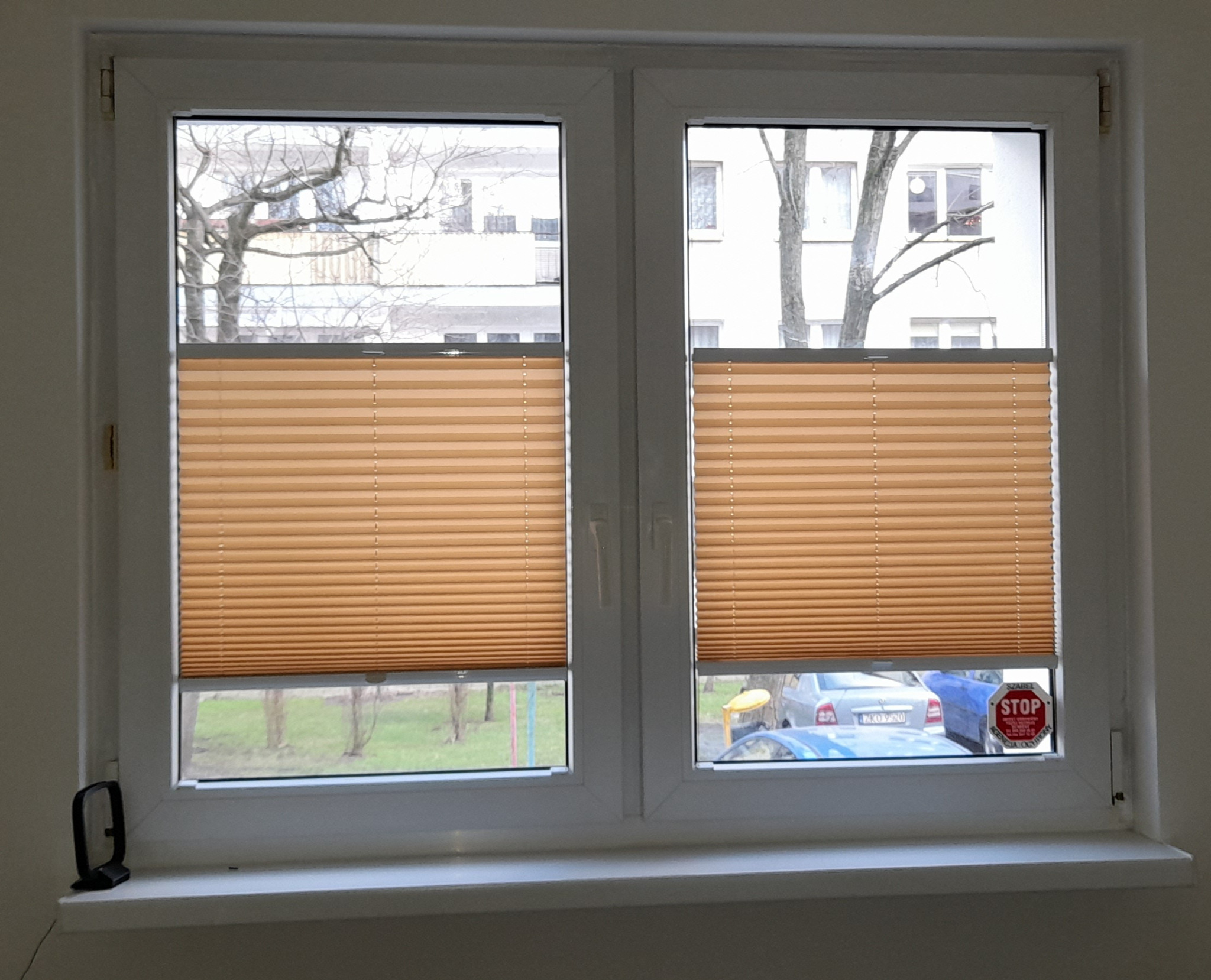 Dwa okna z zamontowanymi plisami w kolorze beżowym, na jednym oknie naklejka 'STOP'. Widok z okna na drzewa i zaparkowane samochody.
